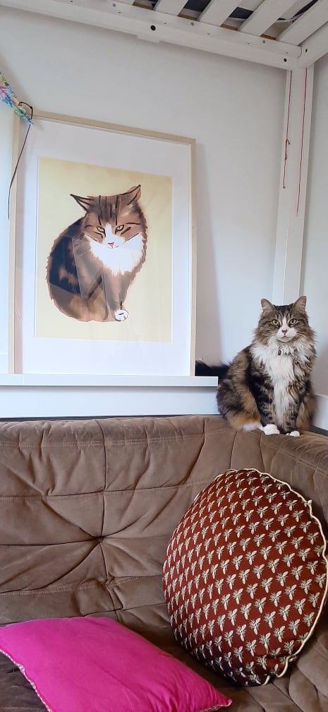 Chatte assise sur le dossier d’un canapé marron devant un portrait d’elle plus que grandeur nature que j’ai dessiné. Elle regarde droit dans l’objectif.