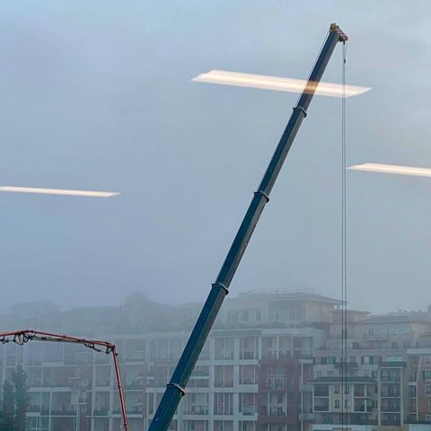 Immeubles et grues sur fond de ciel gris et brumeux, pris à travers une baie vitrée dans laquelle se reflètent des néons au plafond.