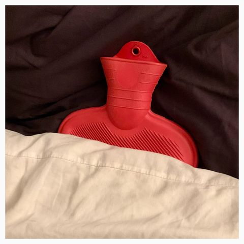 A red water bottle under a white comforter sitting up against a silky black pillow.
