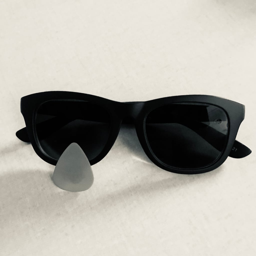 Photo en noir et blanc d’une paire de lunettes de soleil posées sur une table. Un plectre de guitare est posé cul en bas devant un verre des lunettes, pour suggérer une larme.
